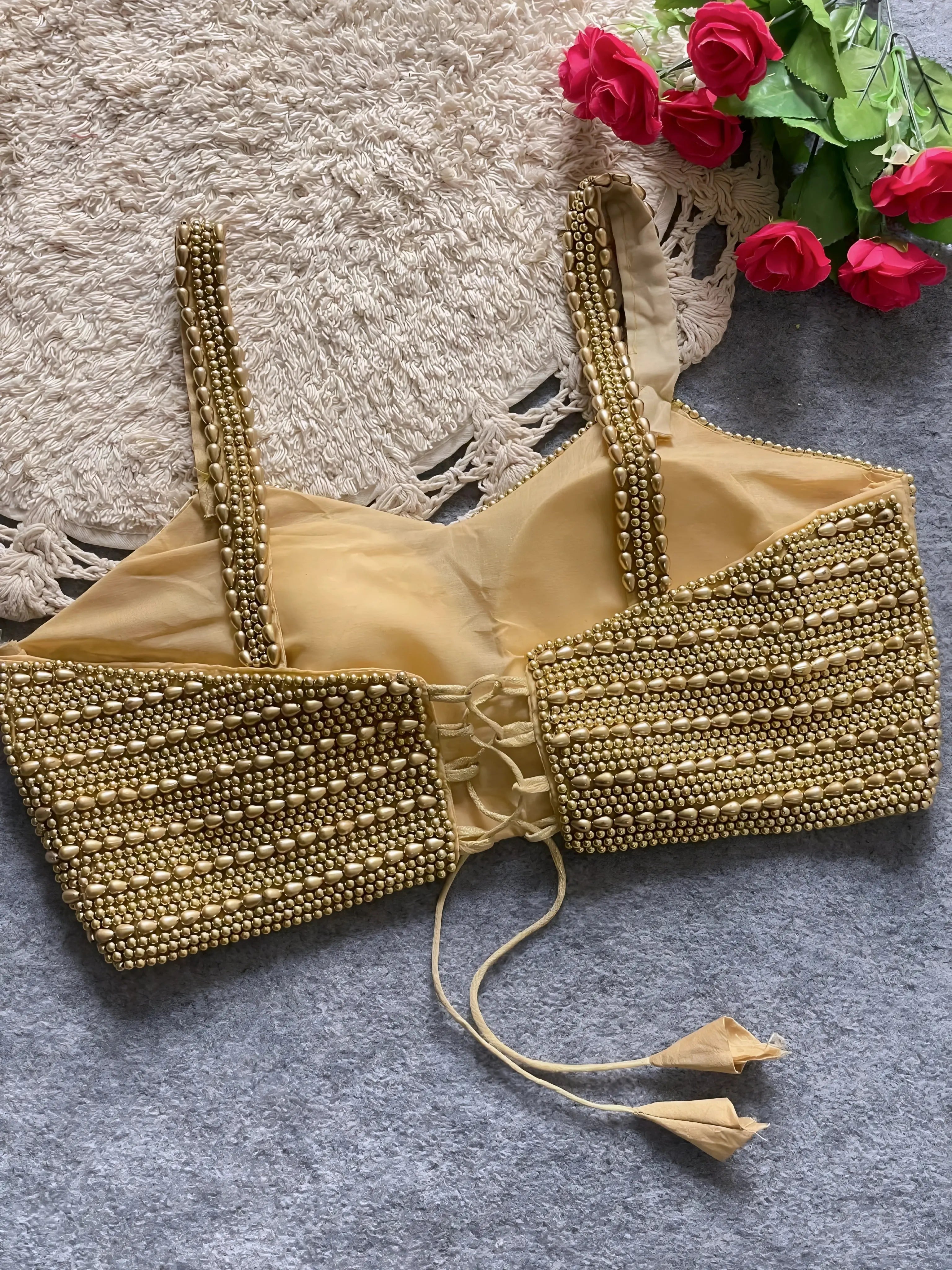 Traditional gold blouse featuring intricate beaded and sequin embellishments in horizontal rows, wide shoulder straps, and a tasseled tie-back detail, displayed flat on a textured grey surface with a braided cream mat and red artificial roses in the corner.